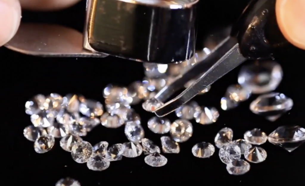 Close-up of several loose diamonds being examined on a black background. A magnifying tool is held over one diamond, and tweezers are positioned to pick it up.