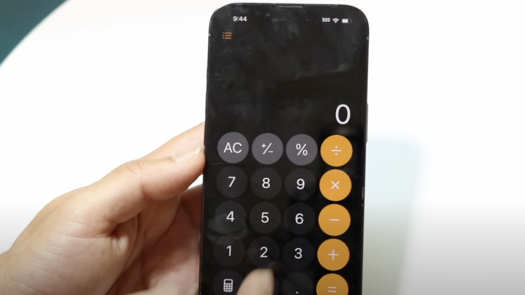 A hand holding a smartphone with a calculator app open. The screen displays large buttons for numbers and basic arithmetic operations like addition, subtraction, multiplication, and division. The calculator shows "0" as the current entry.