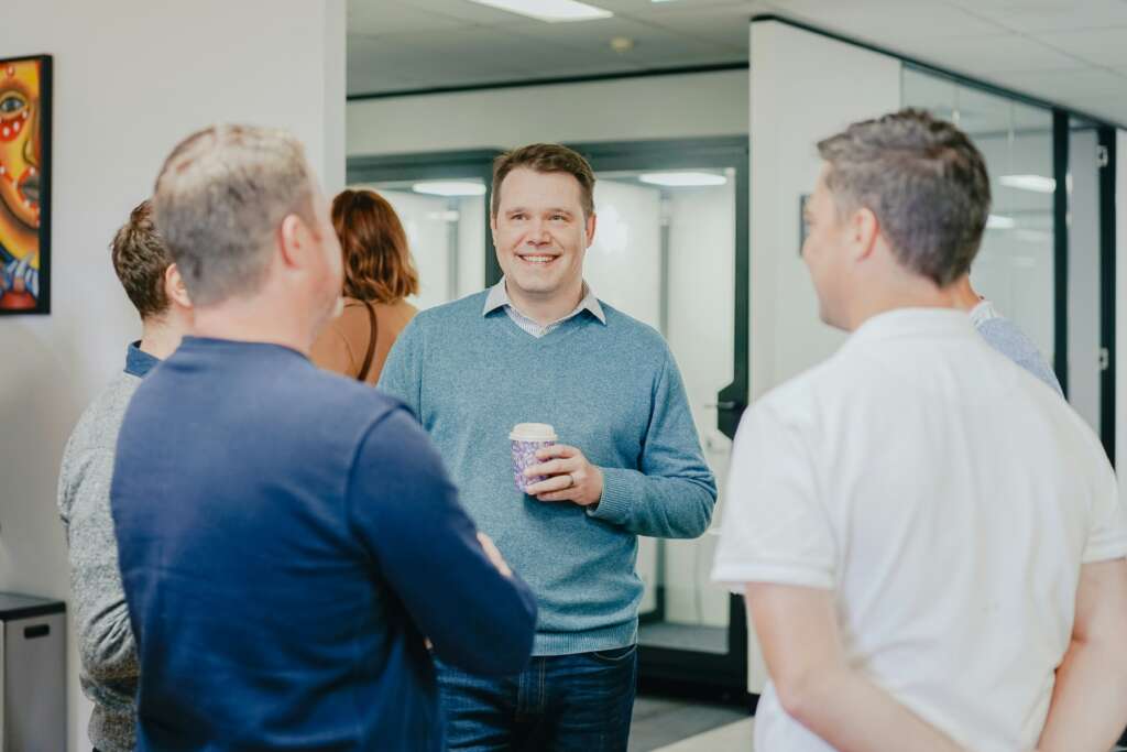 A group of people engaged in conversation in an office setting. A man in the center is smiling and holding a coffee cup. Other people, both men and women, are standing around him. White walls and glass partitions are visible in the background.