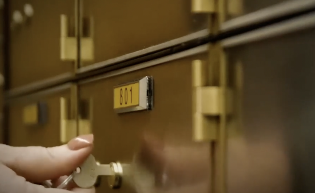 A hand with a key is about to unlock a brass-colored safe deposit box numbered 801. The box is part of a series of similar boxes, each with a small metal plaque and sturdy hinges.