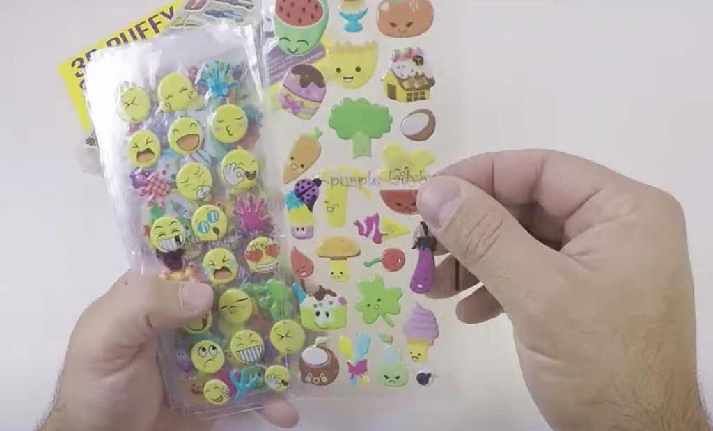 Hands holding puffy sticker sheets; one features emojis with different expressions, and the other has cute food and nature-themed characters like fruits, desserts, a tree, and an eggplant.