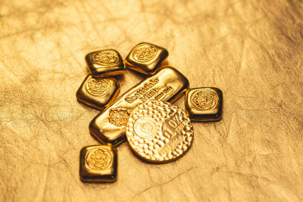 Gold bars and coins arranged on a textured golden surface, displaying ornate designs and inscriptions. The bars are rectangular while the coins are round and embossed, creating a luxurious and shimmering effect.