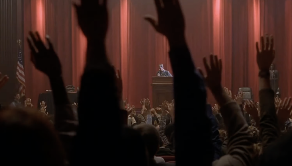 Audience members raise their hands in a dimly lit auditorium with red curtains, listening to a speaker at a podium. An American flag stands on stage, emphasizing a formal event or gathering.