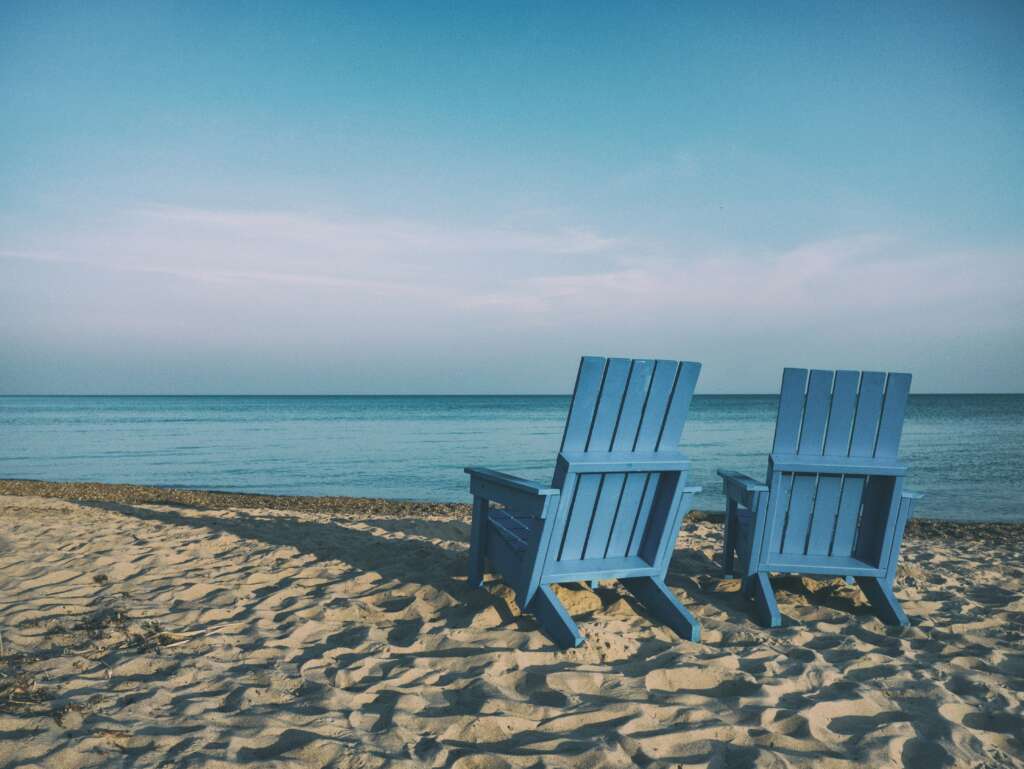 Two blue wooden chairs face the calm ocean on a sandy beach under a clear sky.