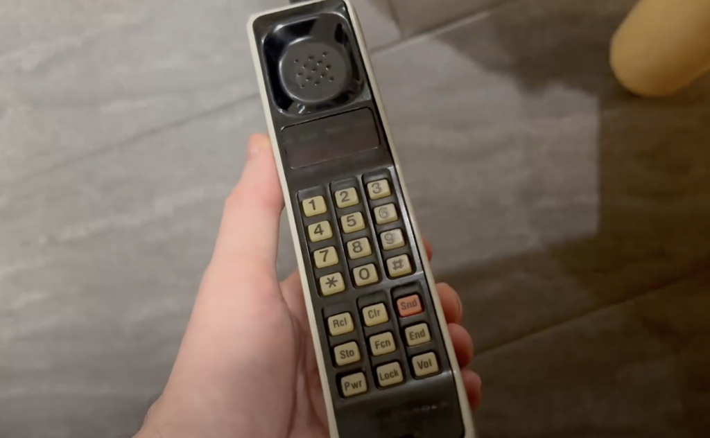 A person holds an old mobile phone with a blocky design and large buttons. The phone has a numeric keypad and a speaker at the top. The background features a gray, tiled surface.