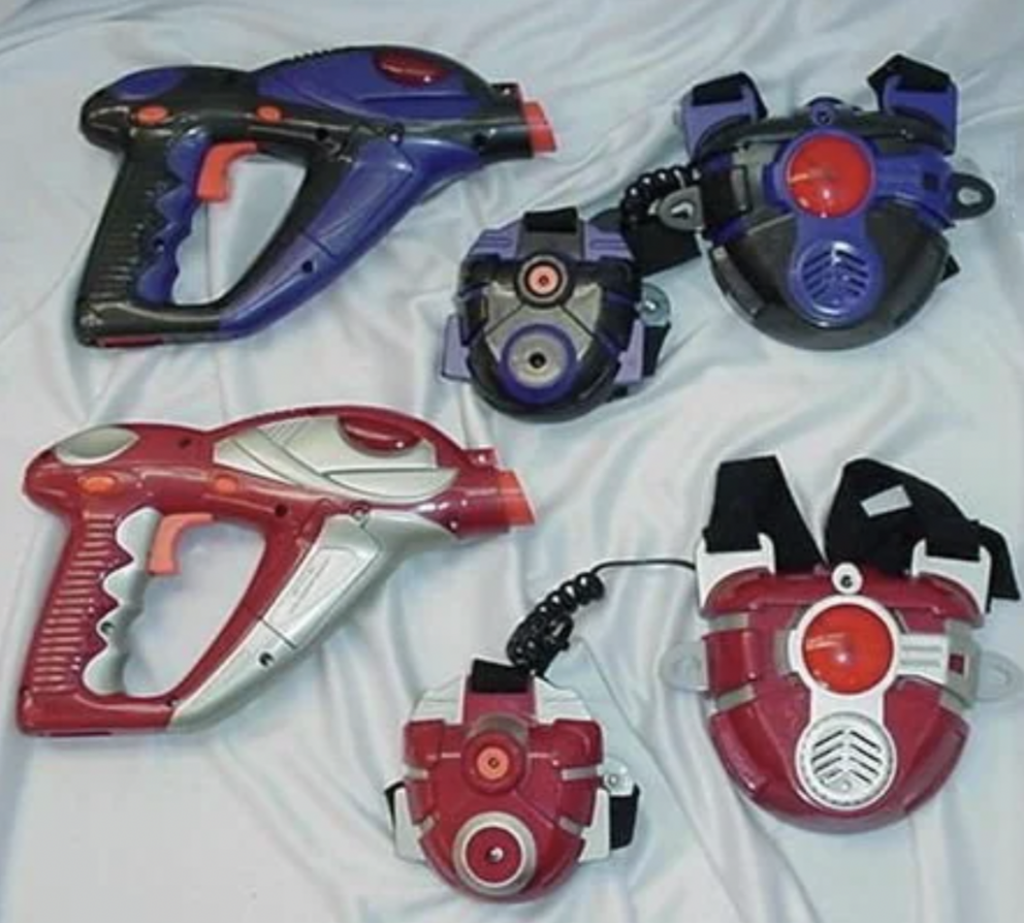 Two toy laser guns and three chest-mounted targets are laid out. The guns are designed in blue and purple, and red and gray. The targets have matching color schemes with circular shapes and strap attachments. They rest on a light fabric surface.