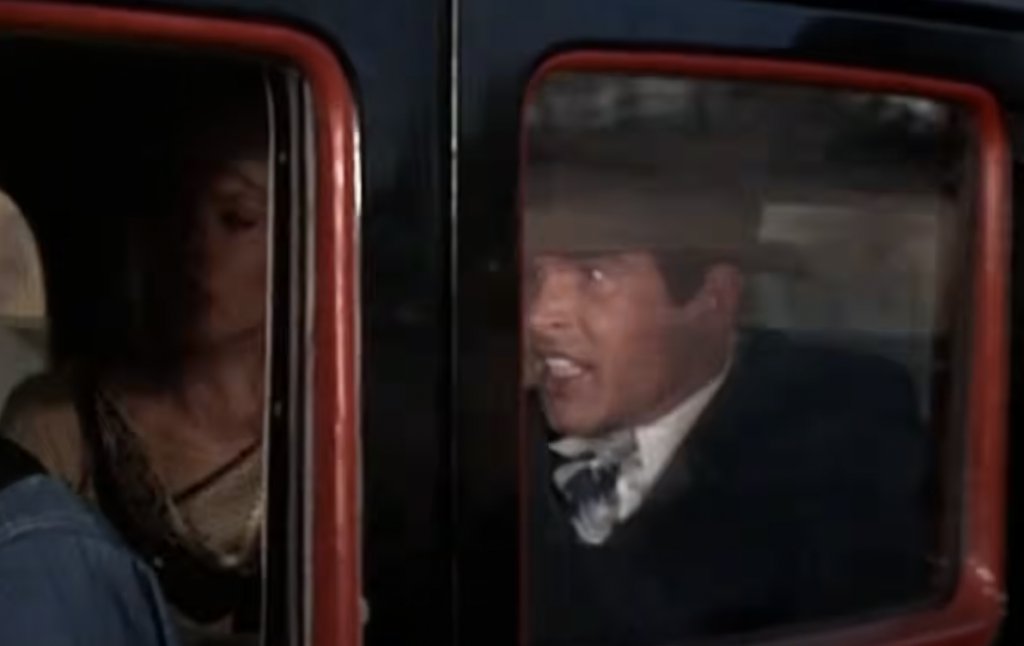 A man wearing a hat is sitting in the back seat of a vintage car, speaking with an expressive look. Beside him, a woman is partially visible. The car door frame is prominent, with red and black accents.