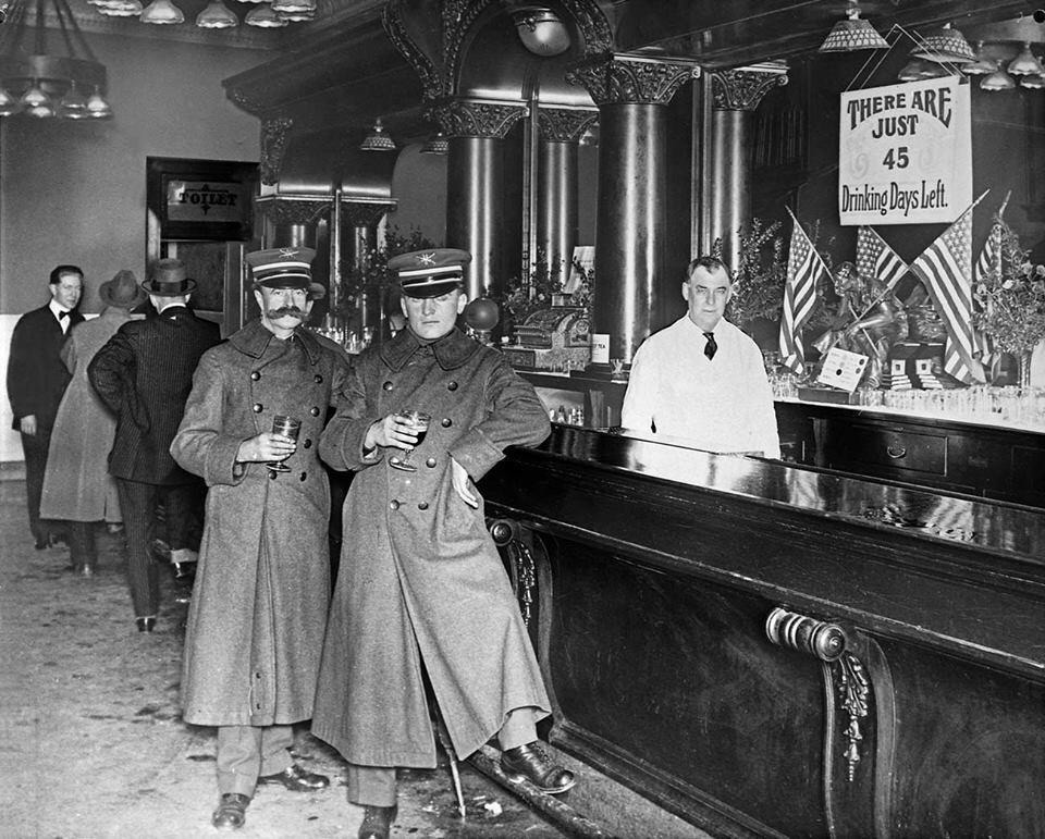 Two men in uniform stand at a bar with drinks in hand. A bartender stands behind the bar. A sign reads, "There are just 45 drinking days left." American flags decorate the bar, and several people are in the background.