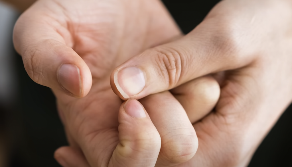 Close-up of a person holding their thumb with the other hand. The focus is on the thumb, showing detailed texture and fingernail. The background is blurred.