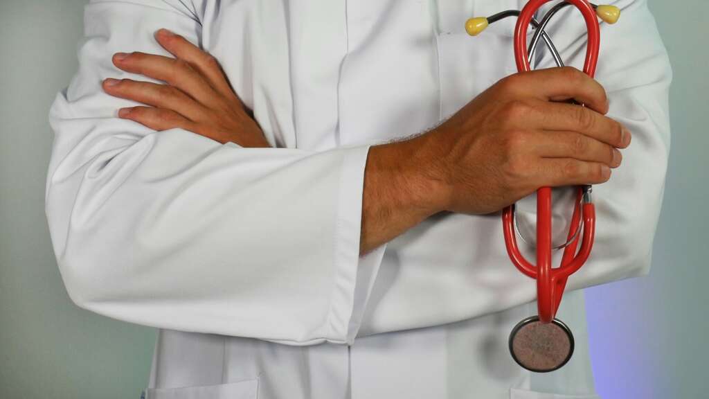 A person in a white lab coat stands with arms crossed, holding a red stethoscope in their right hand. The stethoscope has yellow earpieces and a metal diaphragm. The background is a plain, light color.