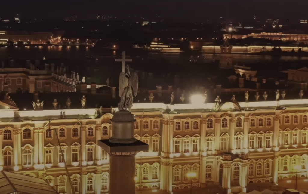 Night view of a grand, illuminated palace with multiple windows, statues, and a prominent angel-topped column in the foreground. The scene overlooks a lit city, with a river visible in the distance.