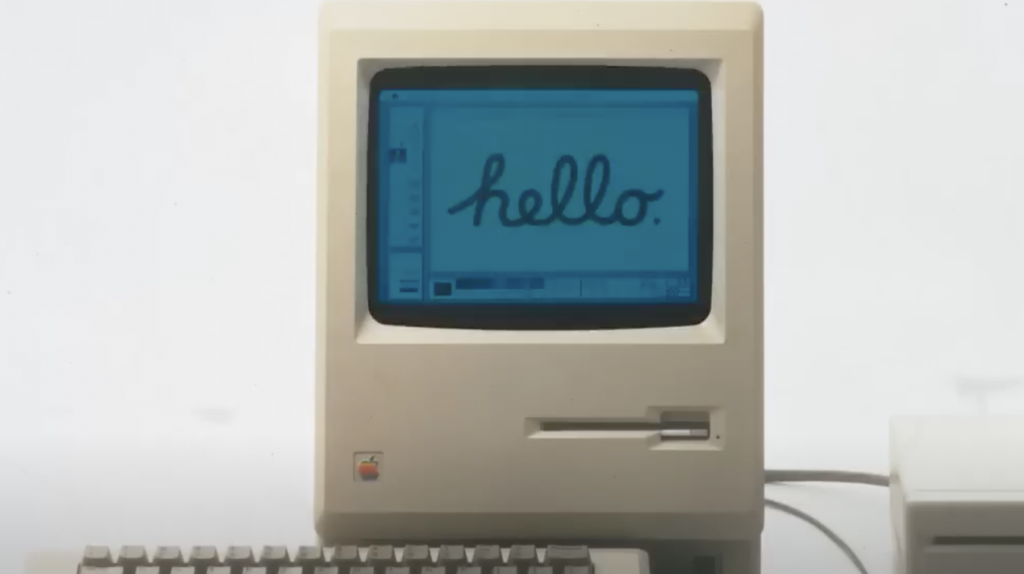 A vintage computer with a beige casing displays the word "hello" in script on its blue screen. A keyboard is visible in front of the monitor.