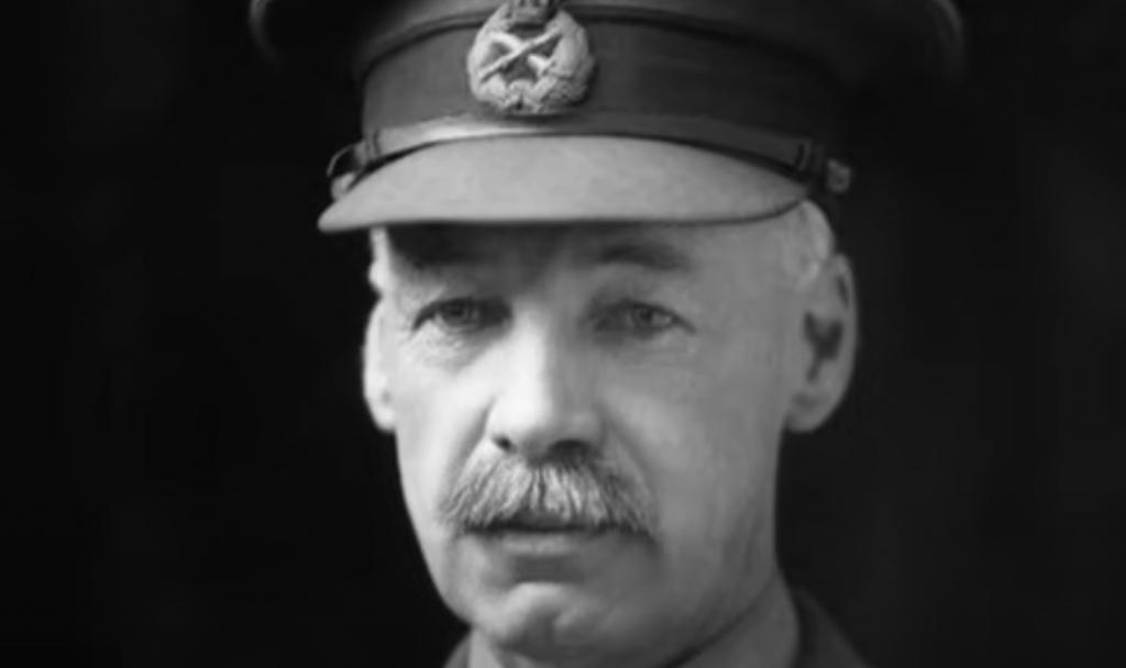 A black and white photo of an older man wearing a military-style hat with an emblem. He has a mustache and is dressed in a uniform, looking directly at the camera with a neutral expression against a dark background.