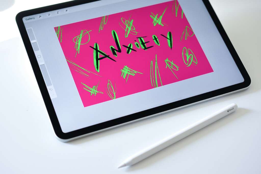 A tablet on a white surface displays the word "Anxiety" in black on a bright pink background with green scribbles. A white stylus lies next to the tablet.