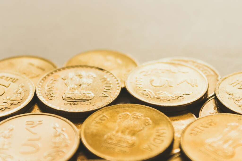 Close-up of Indian coins scattered on a surface. The coins feature various inscriptions and symbols, including the national emblem of India. The surface and the coins have a warm, golden hue.