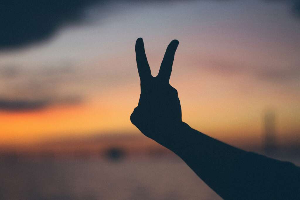 Silhouette of a hand forming a peace sign against a blurred sunset sky and horizon. The sky displays warm hues of orange, pink, and purple, creating a serene and calming atmosphere.