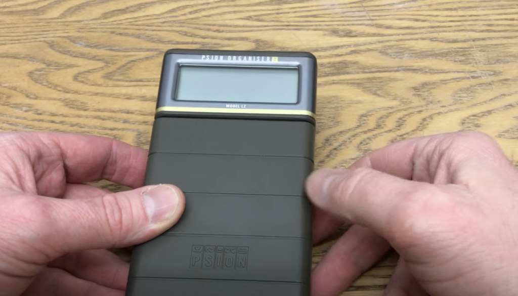 A person holds a Psion Organizer II Model LZ against a wooden surface. The device has a small screen and a dark gray body with a textured grip. The word "PSION" is embossed at the bottom.