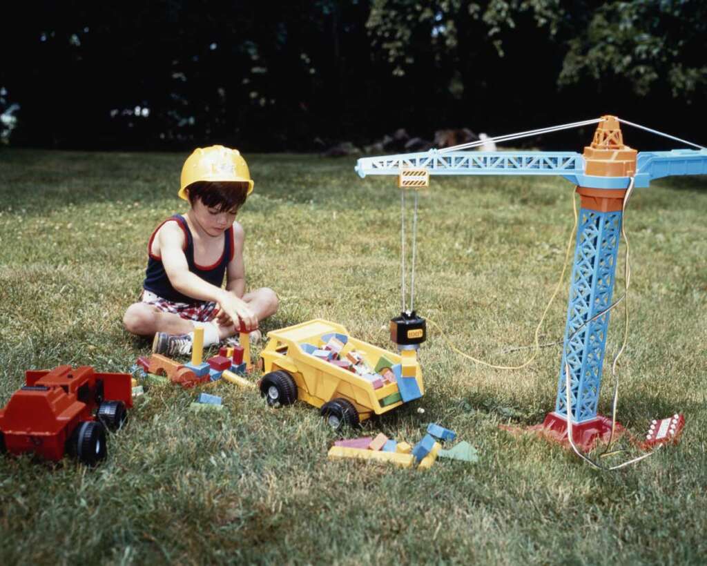 A child wearing a yellow construction helmet plays on grass with colorful building blocks and toy vehicles, including a dump truck, crane, and red trucks. Trees are visible in the background.