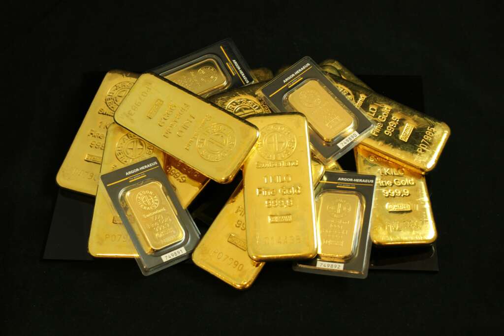 A pile of gold bars of various sizes is stacked against a black background. The bars have engravings and markings, indicating their weight and purity. Some are in protective packaging.