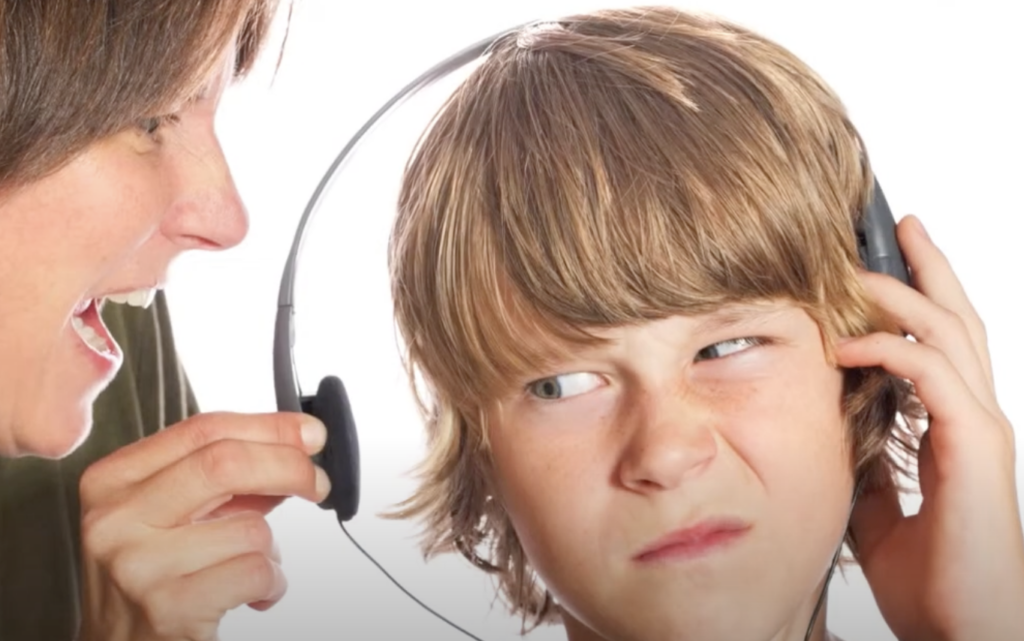 A woman yelling at a boy wearing headphones. The boy is frowning and looking annoyed, seemingly irritated by the loud noise despite the headphones. The background is plain white, emphasizing their expressions and interaction.