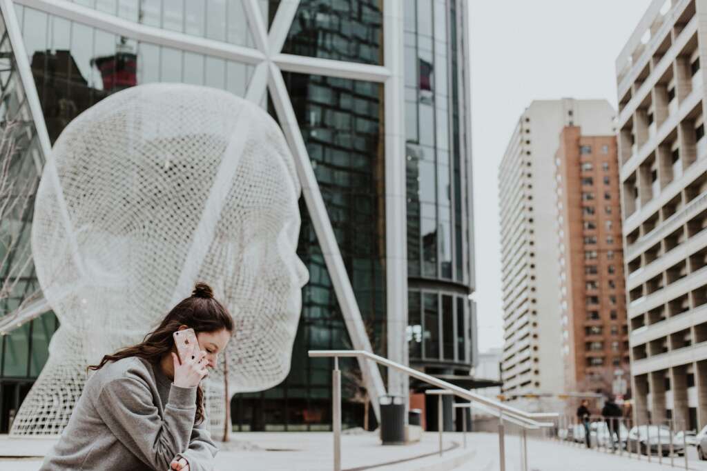 A person sits on steps while talking on a phone in an urban area. Behind them is a large wire mesh sculpture of a head, with modern buildings surrounding the scene. The weather appears overcast.