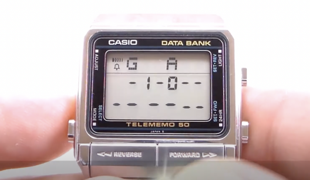 Close-up of a Casio Data Bank Telememo 50 watch with a digital display showing "5 A" and a pattern of dashes. The watch features multiple buttons labeled "Reverse," "Forward," and options for setting time and alarms.