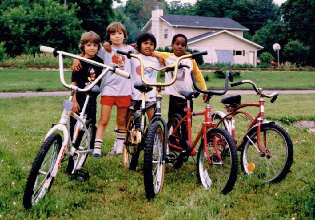 Four children stand in a grassy yard with their arms around each other, smiling. Three bicycles are parked in front of them. A house and trees are visible in the background.