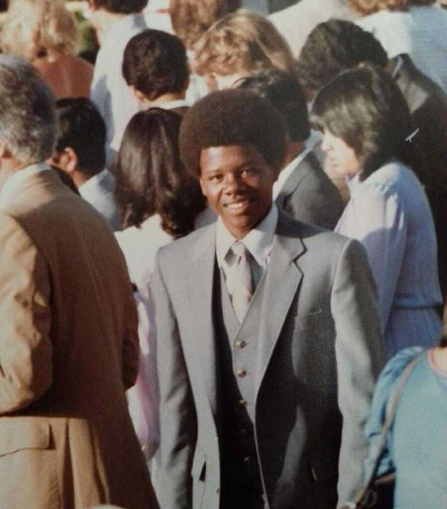 A man in a gray three-piece suit and tie is smiling while walking in a crowd. He has an afro hairstyle and is surrounded by people who are mostly facing away from the camera. The setting appears to be outdoors.