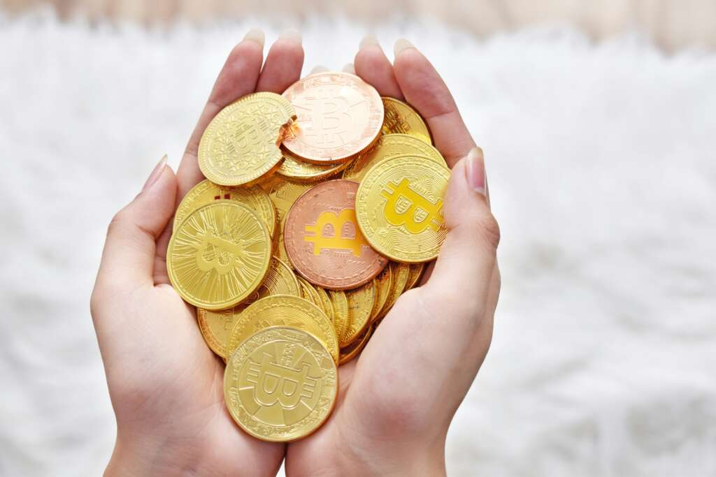 Hands holding a pile of gold and copper-colored coins with a Bitcoin symbol. The coins are displayed against a blurred white background, emphasizing their shiny metallic texture.