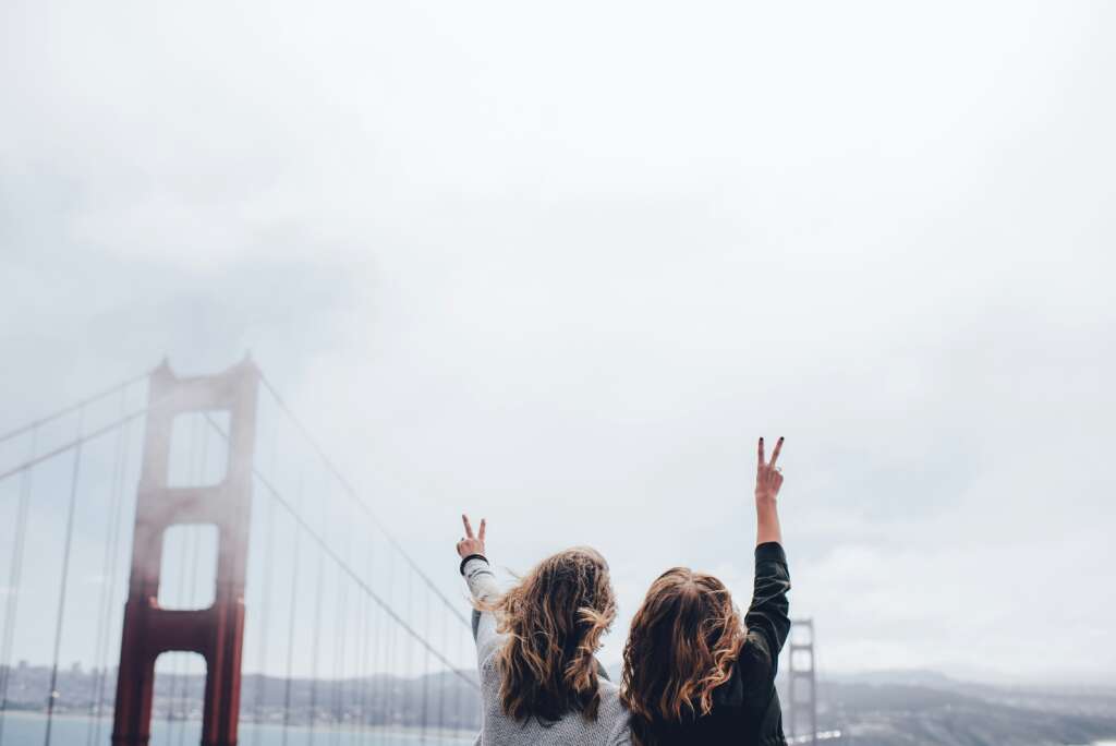 Two people stand with their backs to the camera, facing the Golden Gate Bridge. They raise their hands in a peace sign against a cloudy sky. The iconic red towers of the bridge are partially visible through the mist.