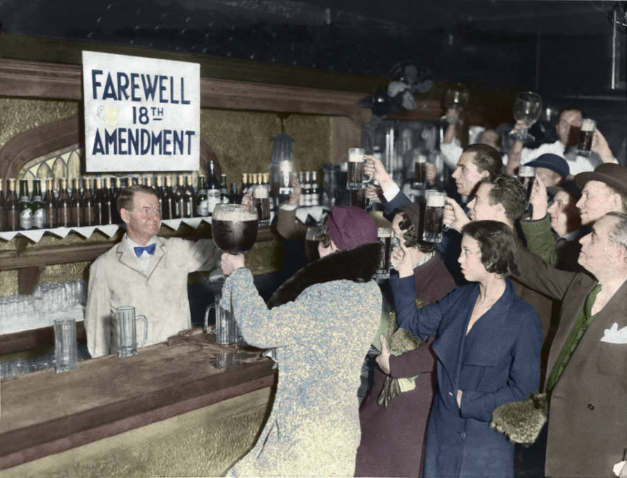 A group of people raise glasses in a bar, celebrating. A sign behind the bartender reads "Farewell 18th Amendment." Bottles line the shelf, and the crowd appears joyful. The image is colored, capturing a historic moment of celebration.