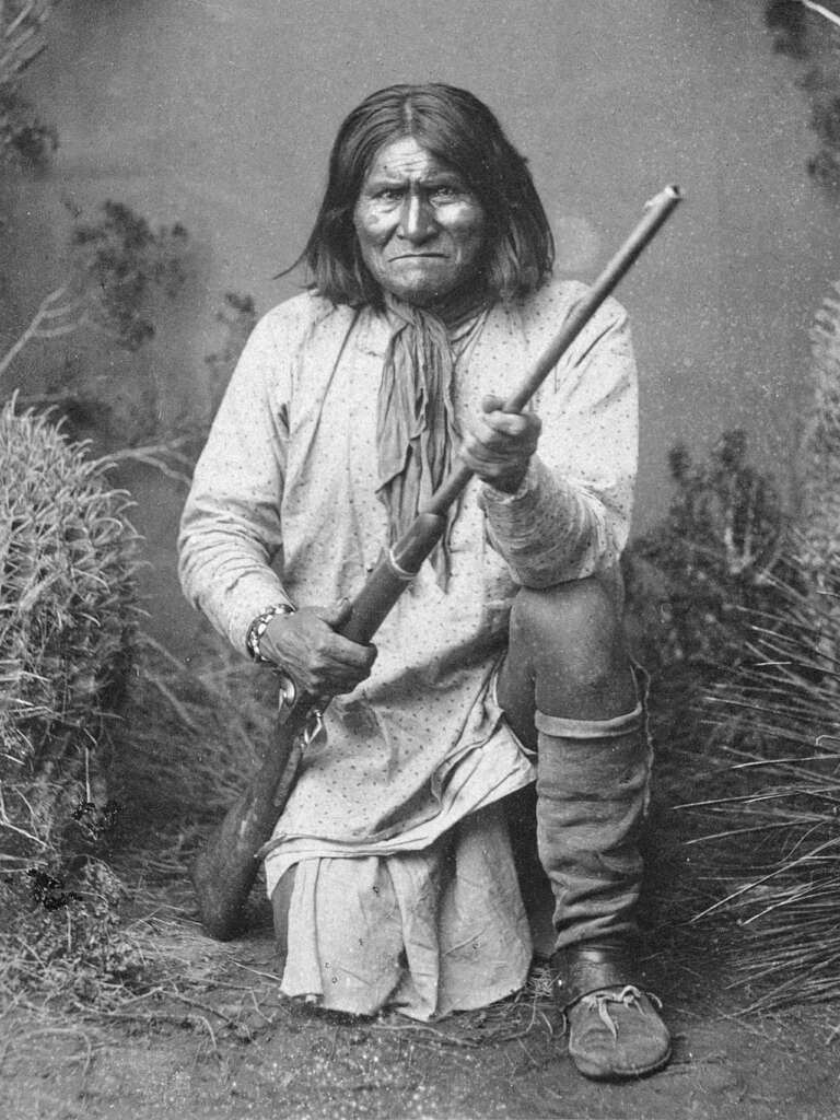 A black and white photo of a person with long hair kneeling in grass, holding a rifle. They wear a long tunic, leggings, moccasins, and a scarf. The background is a studio setting with plants on either side.