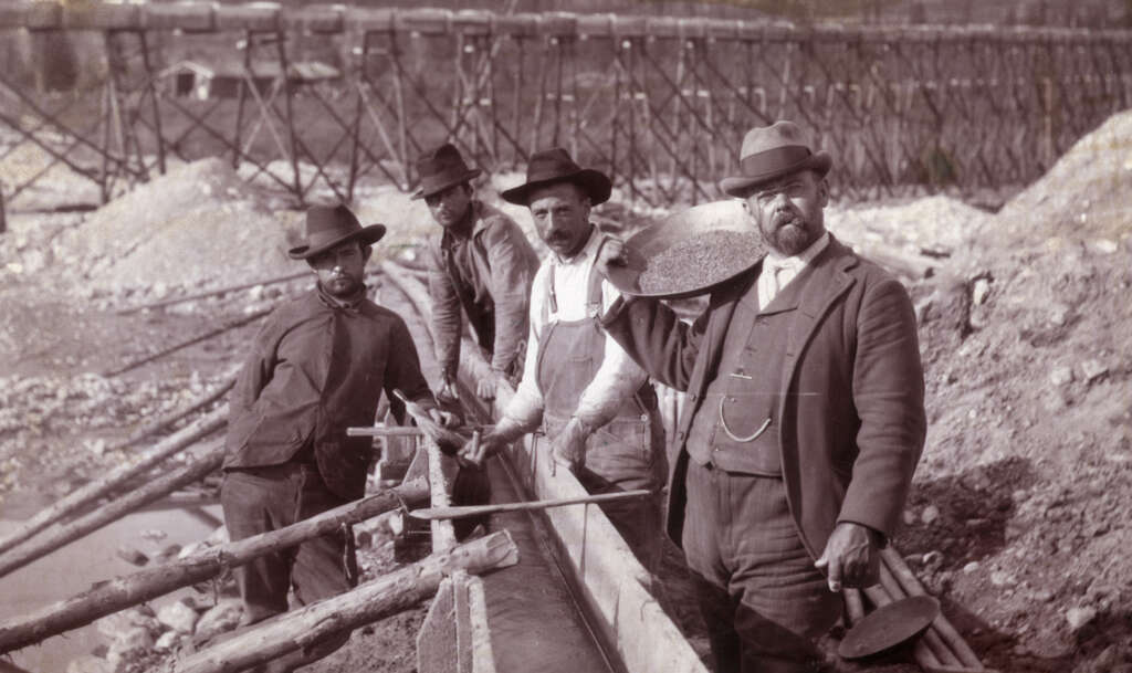 Four men in historical attire stand by a wooden sluice for gold panning. They hold mining tools and pans filled with gravel. A wooden bridge structure is visible in the background. The scene suggests gold mining activities.