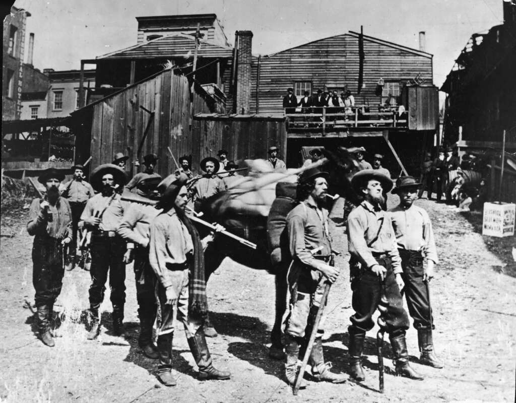 A group of soldiers in uniform stand outdoors in front of a rustic wooden building. Some soldiers hold rifles and wear wide-brimmed hats. A military cannon is visible in the background, and men are seated on a platform above the ground.