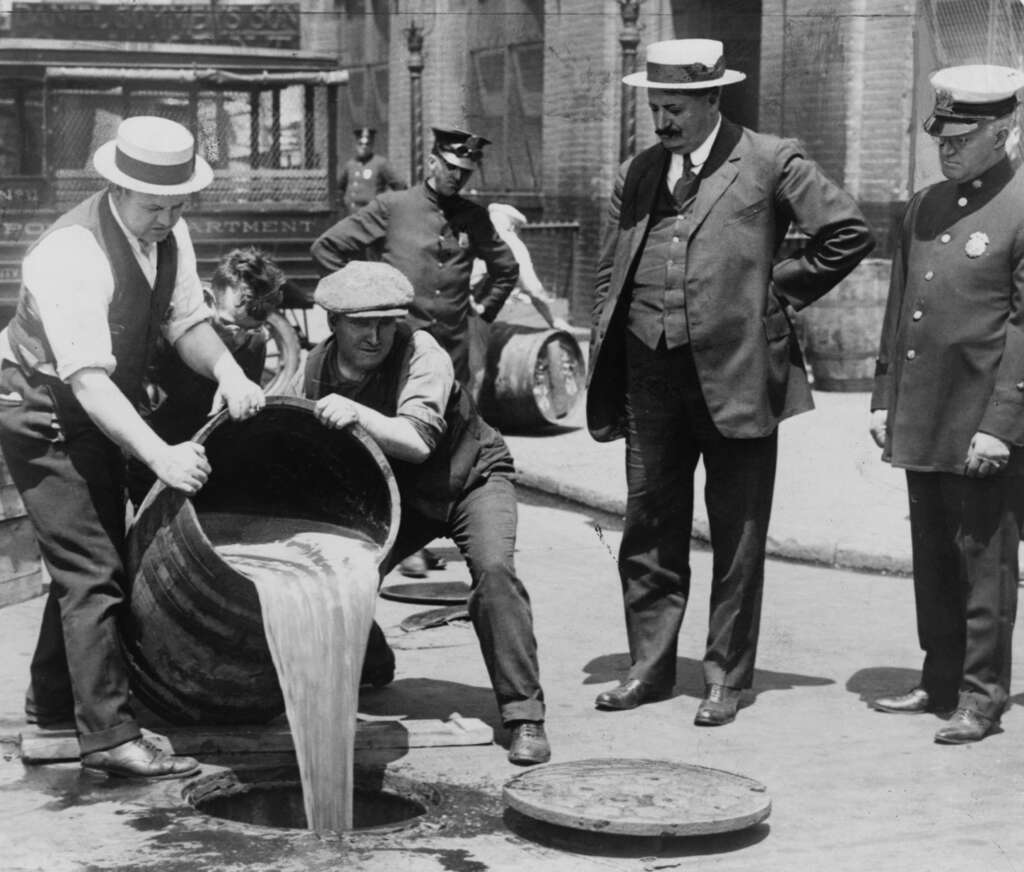 Historical photo of men pouring alcohol into a sewer during Prohibition. Two men handle the barrel while others, including a uniformed officer, look on. A vintage wagon and brick building are visible in the background.