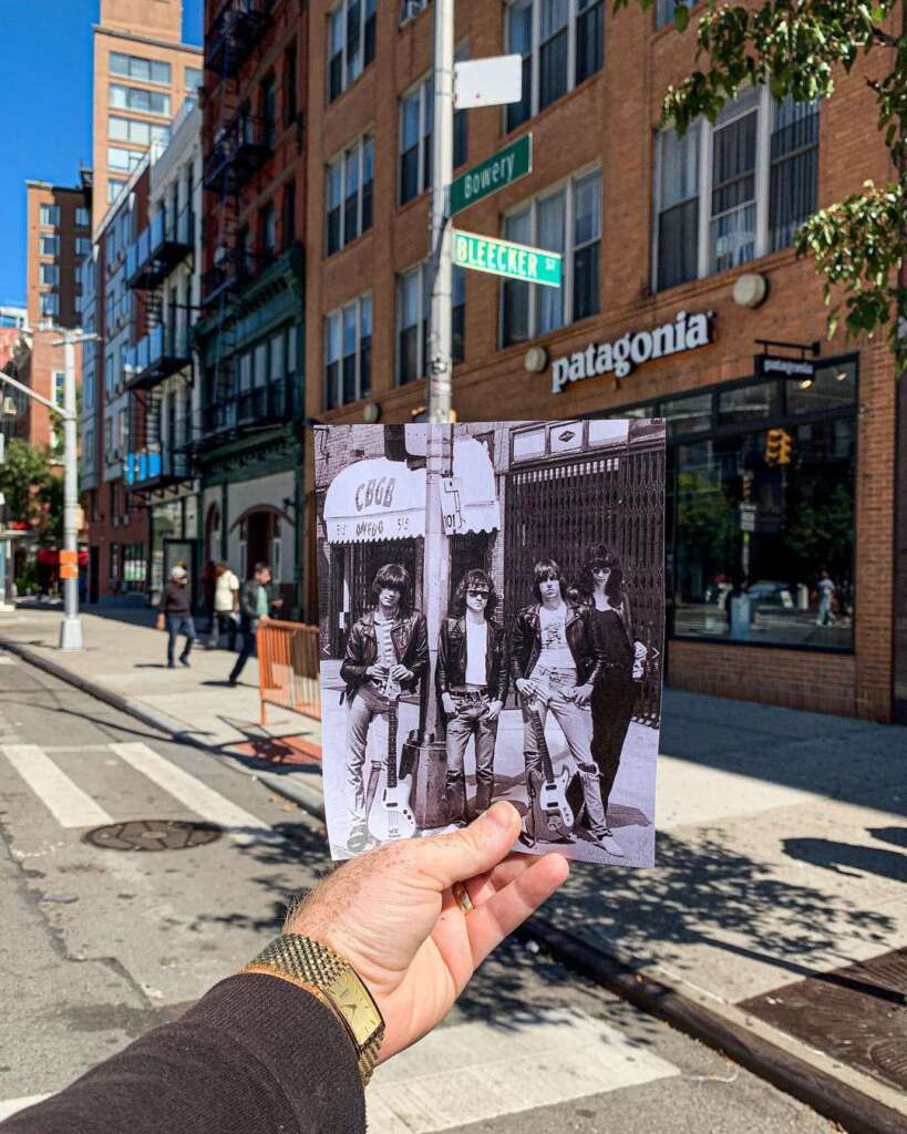 A person holds a vintage black-and-white photo of a band in front of a New York street scene. Behind, a modern-day street corner with a Patagonia store, trees, and urban buildings is visible. The signs read "Bowery" and "Bleecker St.