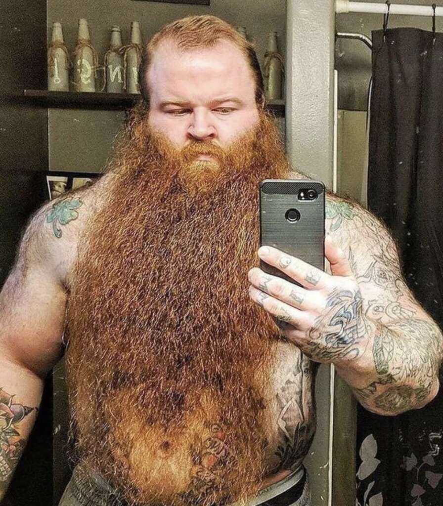 A bearded man with tattoos takes a mirror selfie in a bathroom, holding a smartphone. He has a long, thick beard and is shirtless, showing tattoos on his arms and shoulders. Bottles are visible on a shelf in the background.