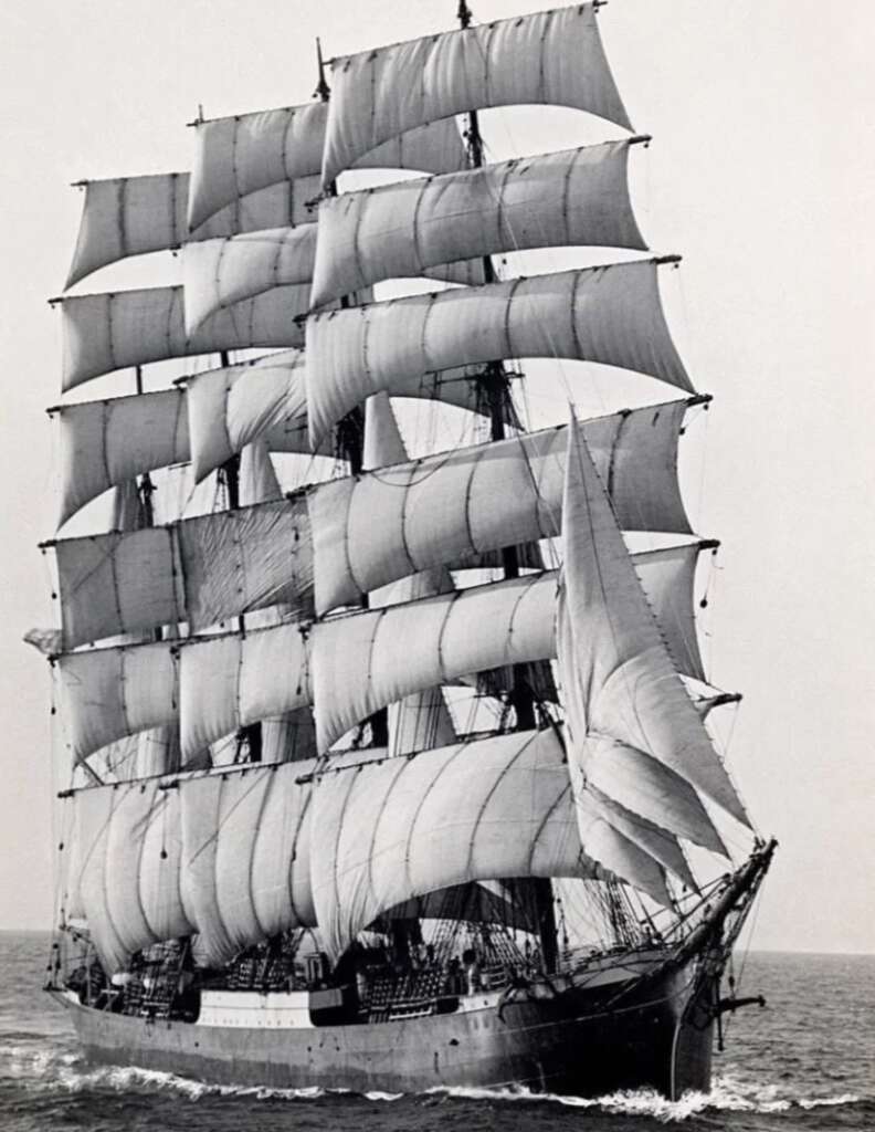 A black-and-white image of a majestic, multi-mast sailing ship with full sails billowing. The ship is navigating through the ocean, with waves at the bow. The sky is clear, highlighting the intricate rigging and tall masts.