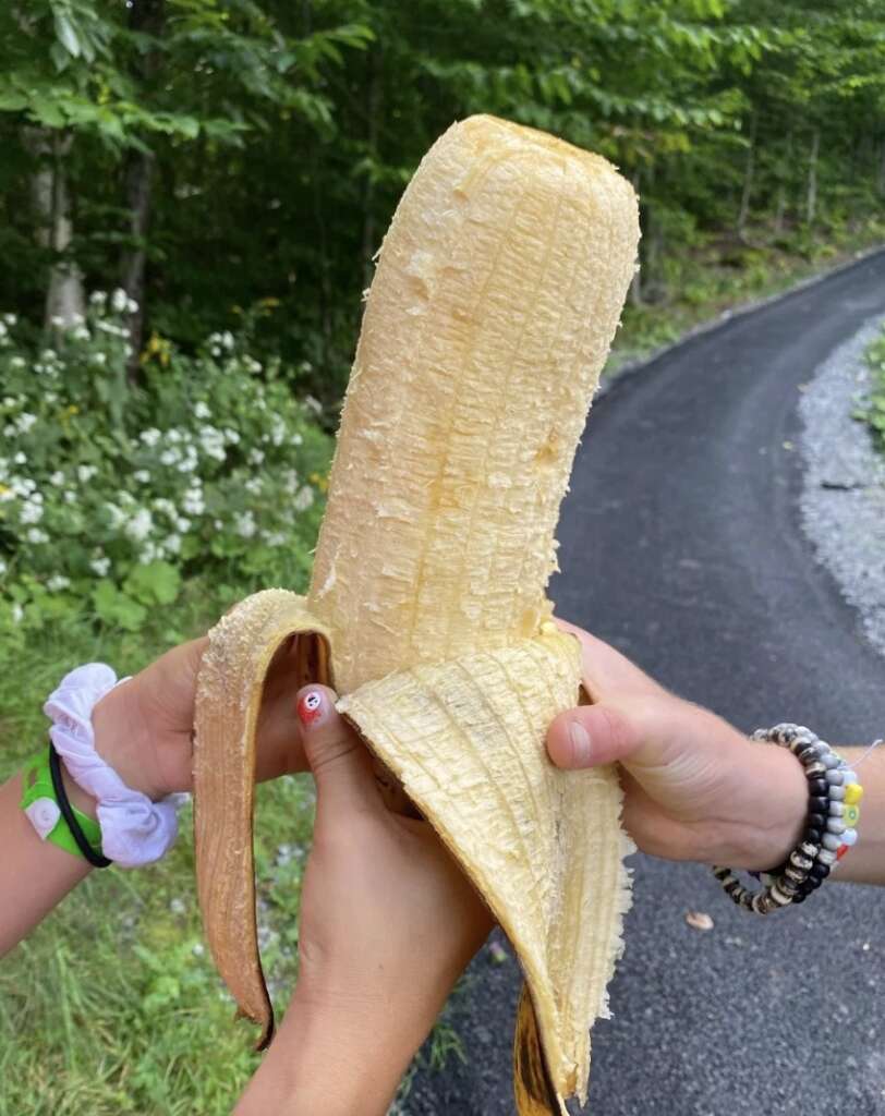 Two hands holding a peeled banana in a garden setting, with greenery and a paved path in the background. One hand is wearing bracelets and the other has a scrunchie.