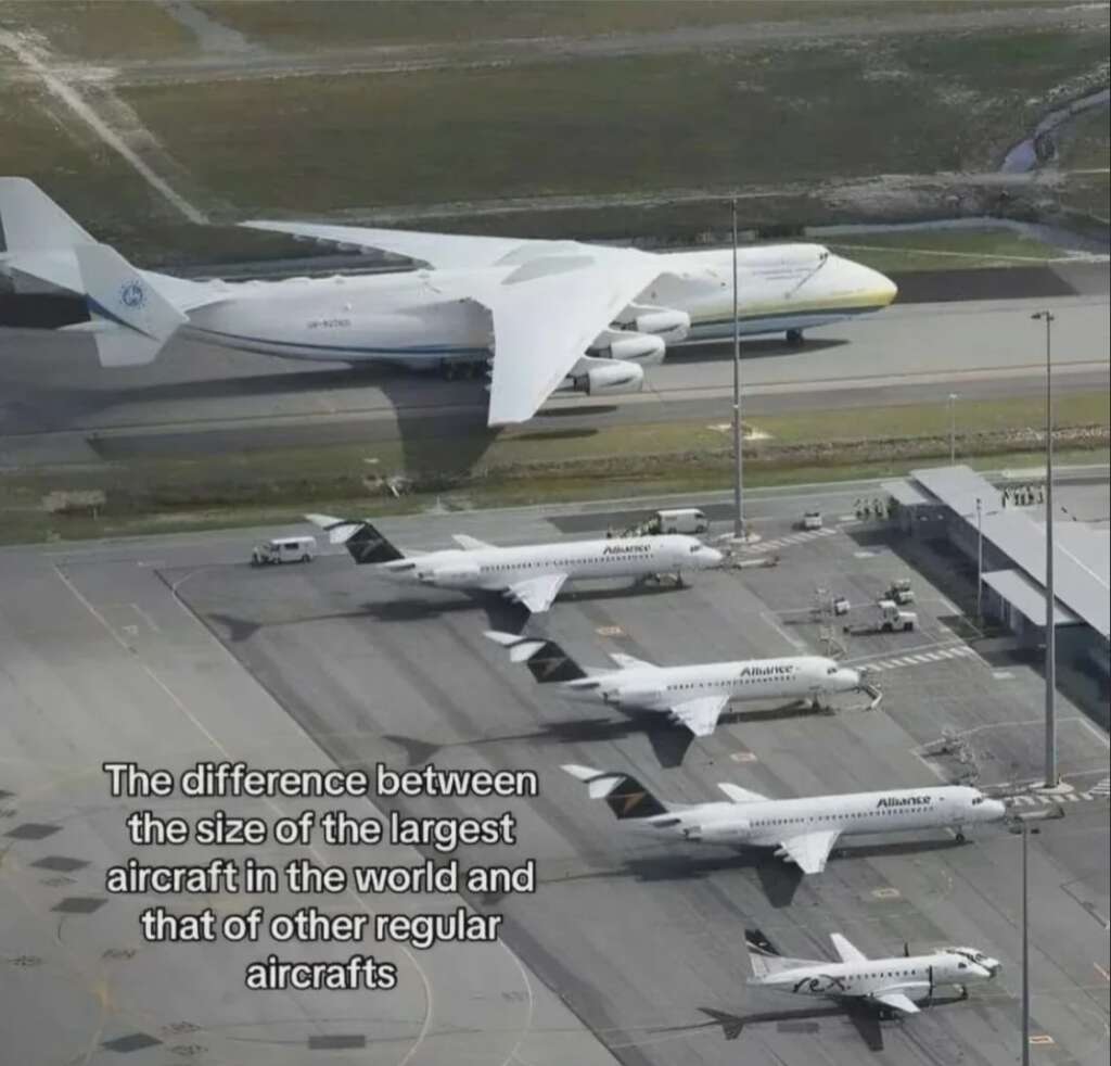 A large aircraft, significantly bigger than others nearby, is parked on a tarmac next to several smaller planes. The image, captured from above, highlights the size difference. Text on image reads: "The difference between the size of the largest aircraft in the world and that of other regular aircrafts.