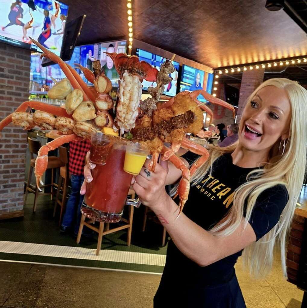 A woman with long blonde hair holds a large Bloody Mary topped with an extravagant seafood and vegetable garnish, including crab legs and fried items. The background is a lively bar with people and TV screens.