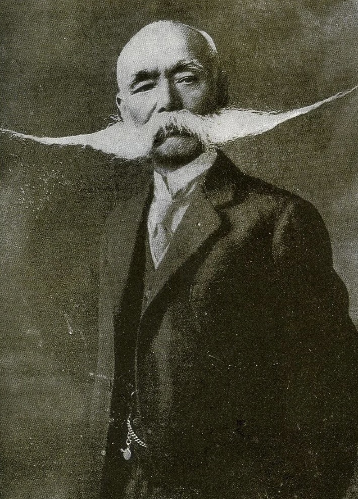 A vintage black and white photo of an older man with a notable, long, and waxed mustache. He is wearing a three-piece suit with a tie, standing against a textured backdrop. His expression is serious and poised.