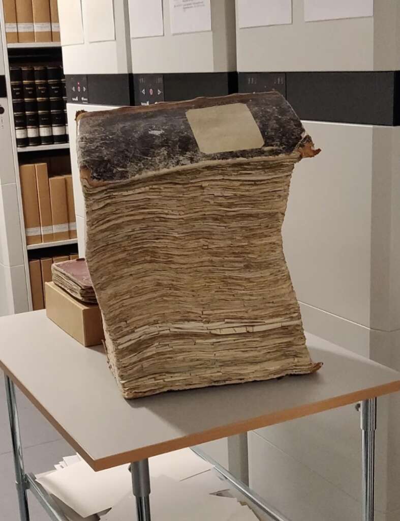 A large, antique book with a worn spine and yellowed pages sits upright on a table in a library or archive setting. Nearby shelves hold additional books and documents.