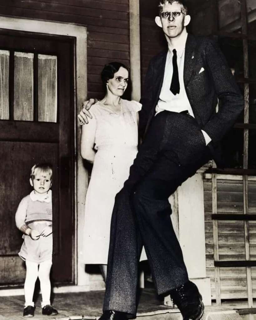 A tall man in a suit stands with his arm on a woman in a dress. A child in shorts and a shirt stands nearby. They are posed in front of a building with a wooden door and steps.