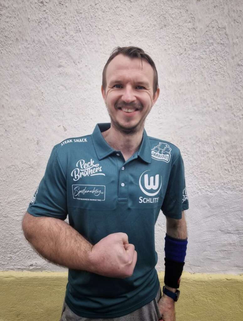 A smiling man in a teal polo shirt with various logos, standing against a textured white wall. He has short hair and a wristband, with one fist raised in a friendly gesture.