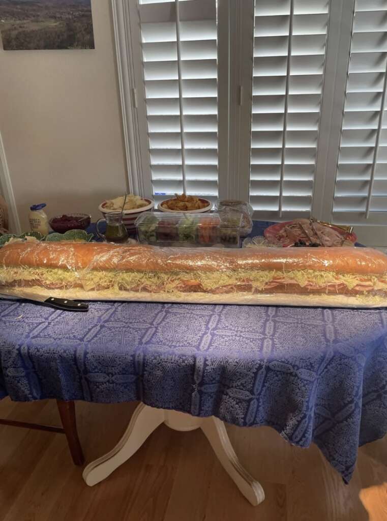 A long sandwich on a blue tablecloth, surrounded by various dishes, including salads and jars, is placed near a window with white shutters. A knife is on the table beside the sandwich.