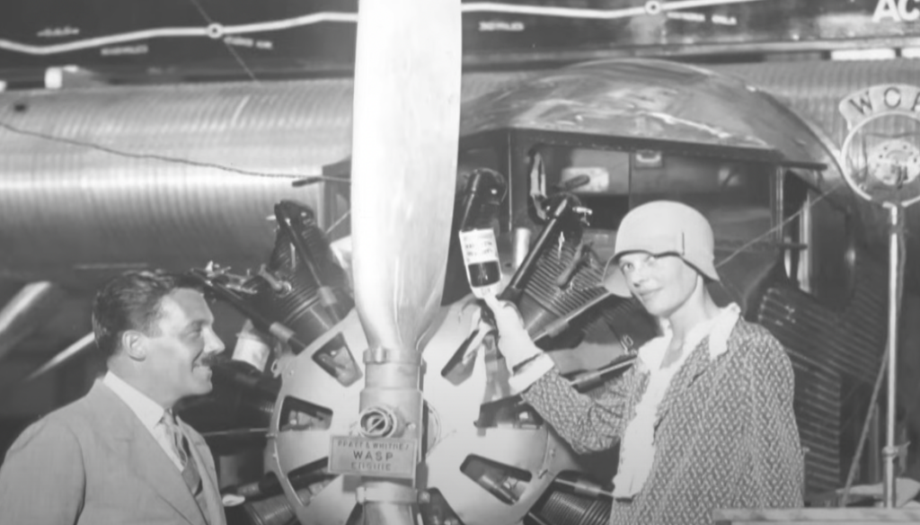 A black and white photo shows a woman in a cloche hat and dress christening a propeller plane with a bottle. A man in a suit stands nearby. The plane has a visible radial engine and a sign with "WORLD WIDE" in the background.