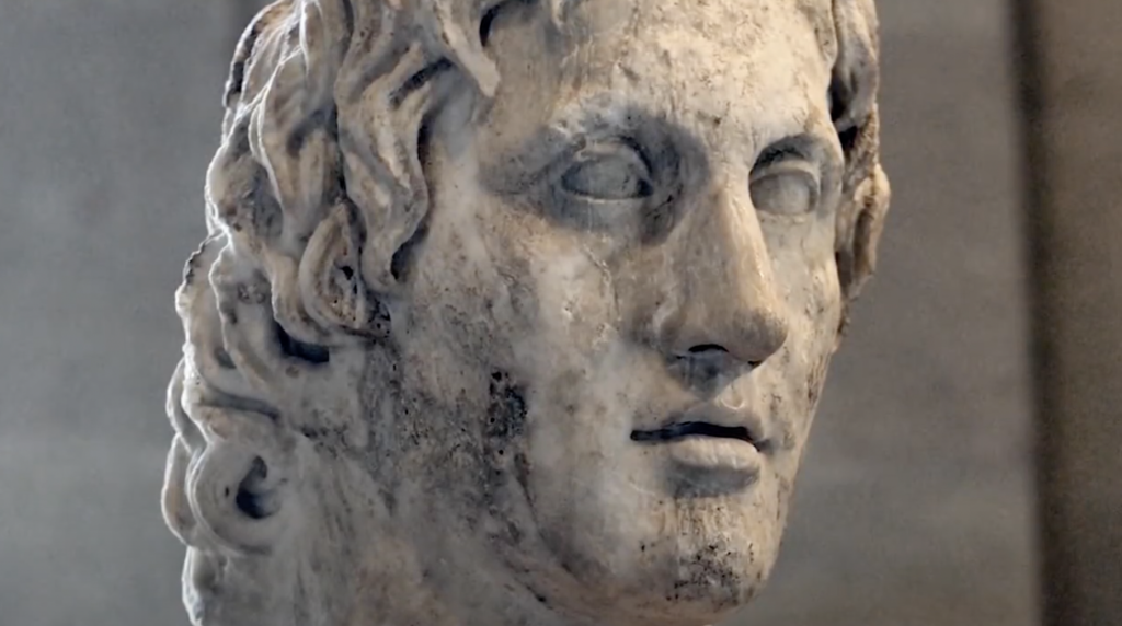 Close-up of an ancient marble bust depicting a man with wavy hair. The sculpture has a weathered texture, and the subject's expression is serene and contemplative. The background is a neutral gray.