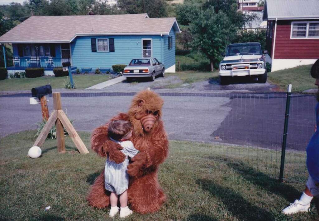 A child in a striped outfit hugs someone wearing a brown furry costume resembling a fictional alien character, on a grassy area in a suburban neighborhood. Two houses and parked cars are in the background.