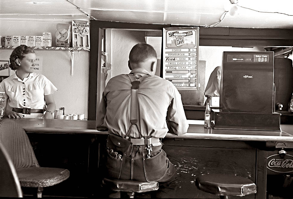 A black-and-white photo of a diner scene. A man sits at the counter wearing a uniform, his back facing the camera. A woman stands behind the counter, looking towards him. The menu above lists items like coffee and hamburgers. An old cash register is visible.